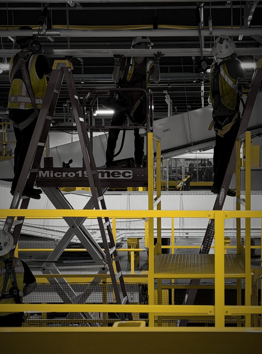 Three installers in hard hats and high-visibility vests working overhead from ladders and a scissor lift in a warehouse facility